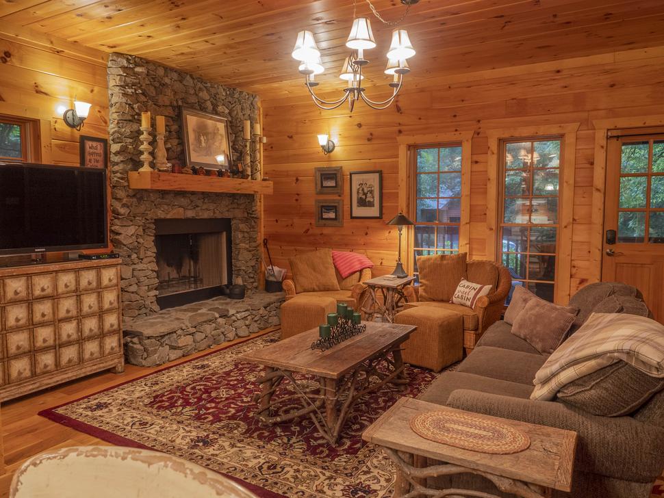 A cozy, wood-walled living room with a stone fireplace, TV, and comfortable seating, illuminated by a chandelier and natural light from large windows.