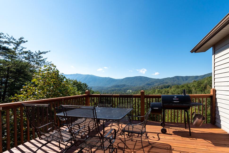 A wooden deck with a table, chairs, and a grill, overlooking a scenic view of mountains and lush greenery under a clear blue sky.