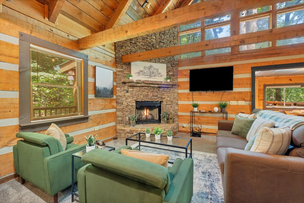 A cozy wooden cabin living room with a stone fireplace, green armchairs, a brown sofa, a flat-screen TV, and large windows offering a view of the forest outside.